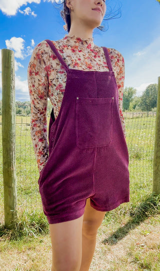 Burgundy corduroy short overalls layered over a sheer floral mock neck top in warm autumn hues.